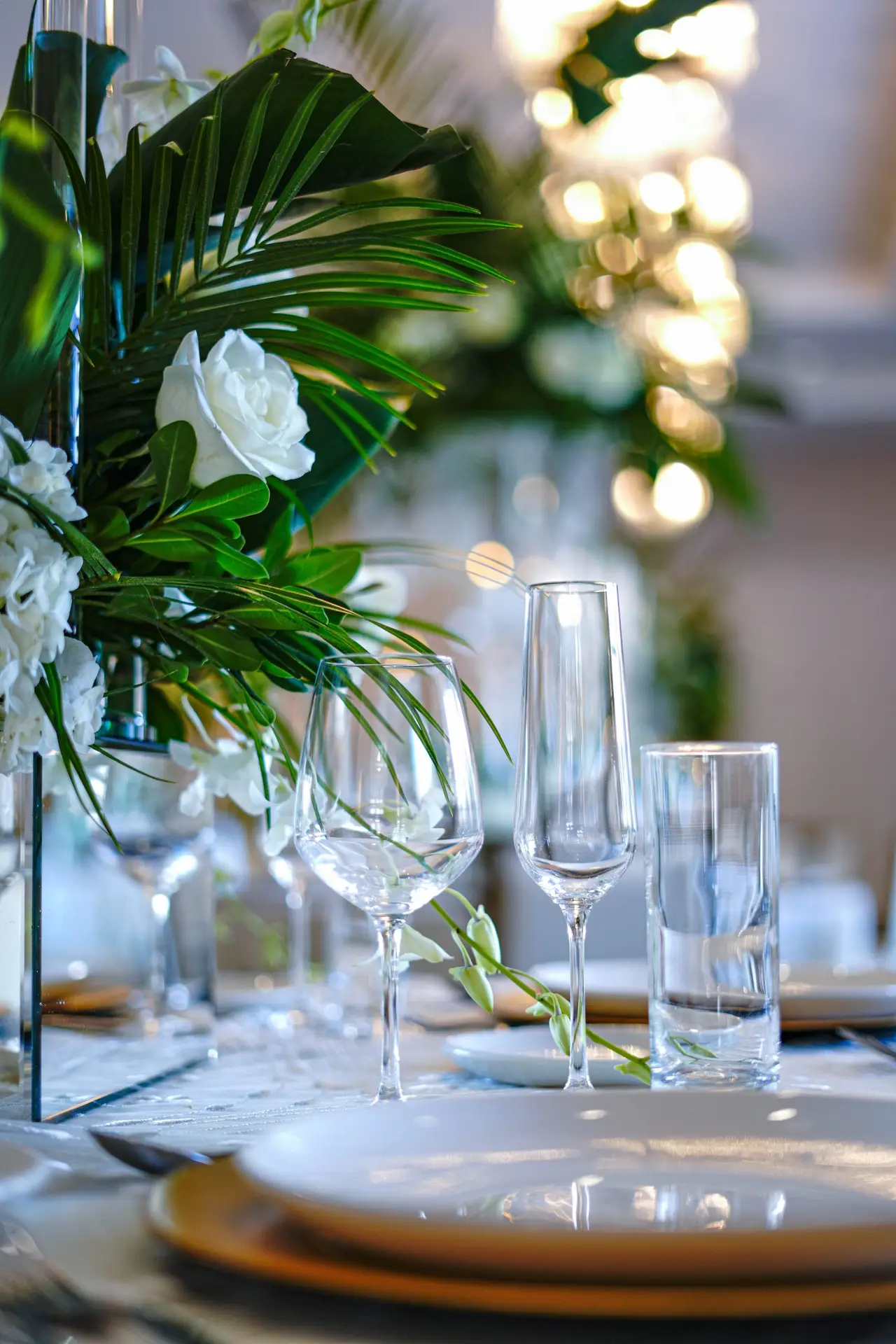 Event table set with a wine glass, champagne flute, and water glass beside a centerpiece of white roses, hydrangeas, and palm fronds