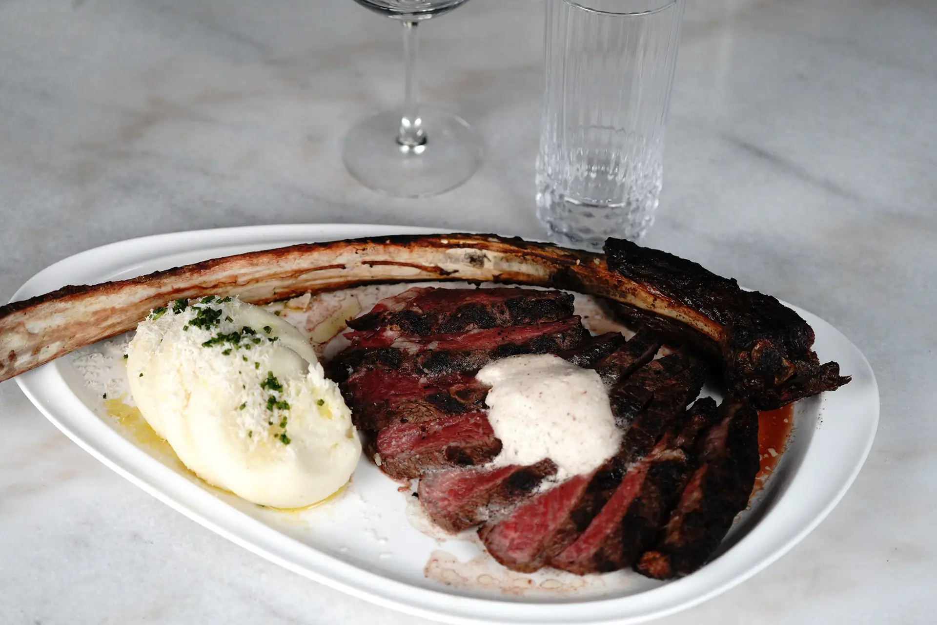 Sliced tomahawk ribeye with whipped potatoes and a creamy sauce on a white platter, with wine and water glasses on marble