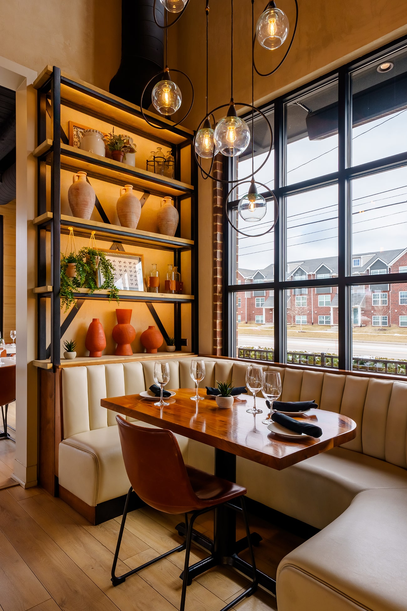 Corner restaurant booth with cream tufted seating and a set wood table beside tall windows and globe pendant lights