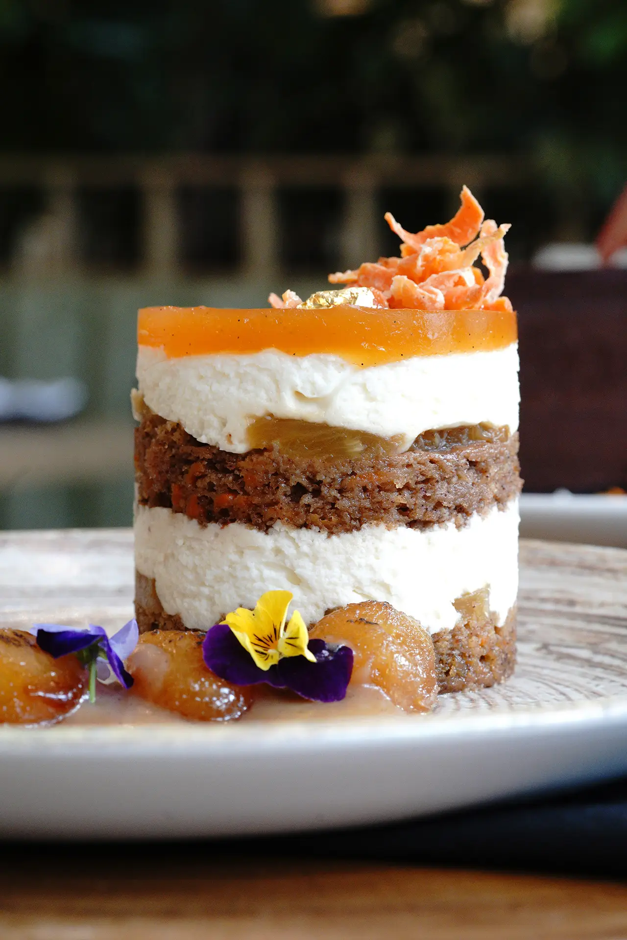 Layered carrot cake with cream cheese mousse, a glossy apricot glaze, candied carrot curls, and gold leaf, plated with caramelized fruit and purple and yellow pansies